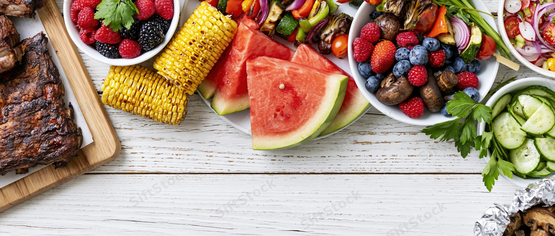 Summer barbecue spread with grilled ribs, corn, watermelon, berries and vegetables arranged along white wooden table edge