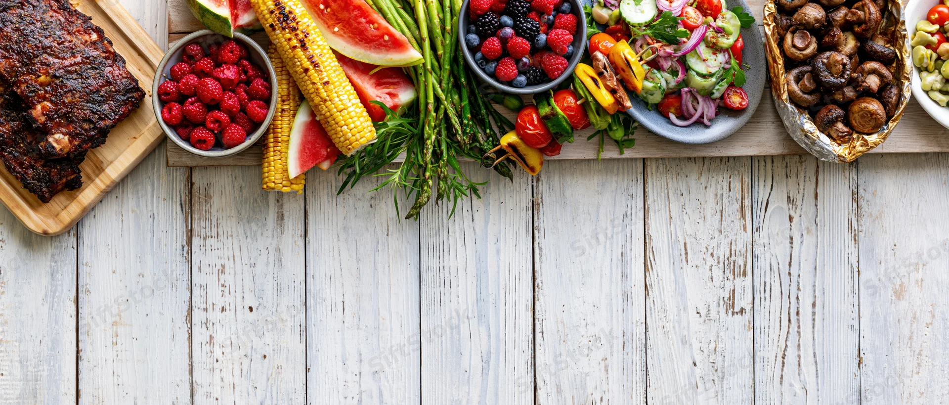 Summer barbecue spread with grilled ribs, corn, watermelon, berries, mushrooms, and vegetables arranged along the left edge of a white wooden table