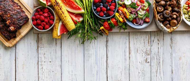 Summer barbecue spread with grilled ribs, corn, watermelon, berries, mushrooms, and vegetables arranged along the left edge of a white wooden table
