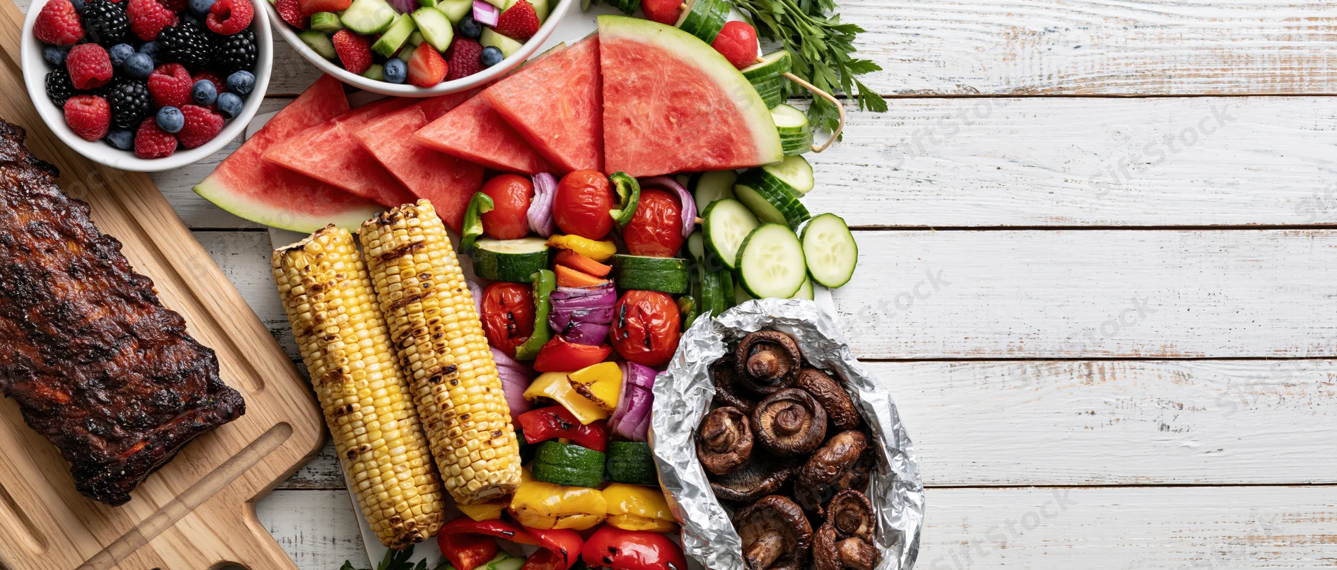 Summer barbecue spread with grilled ribs, corn, watermelon, vegetables and berries arranged along left edge of white wooden table