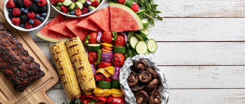 Summer barbecue spread with grilled ribs, corn, watermelon, vegetables and berries arranged along left edge of white wooden table