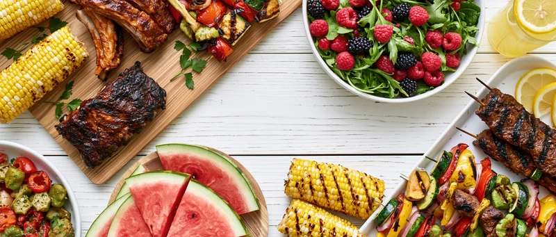 Summer barbecue spread with grilled ribs, corn, watermelon, salad and skewers arranged on a white wooden table