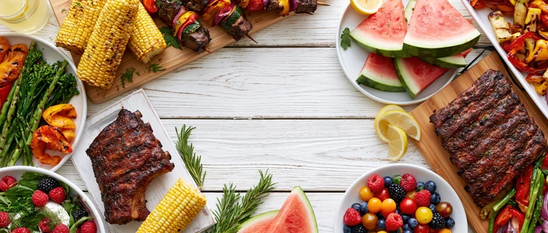 Summer barbecue spread arranged as double border frame on white wooden table with grilled ribs, corn, watermelon, vegetables and fruit