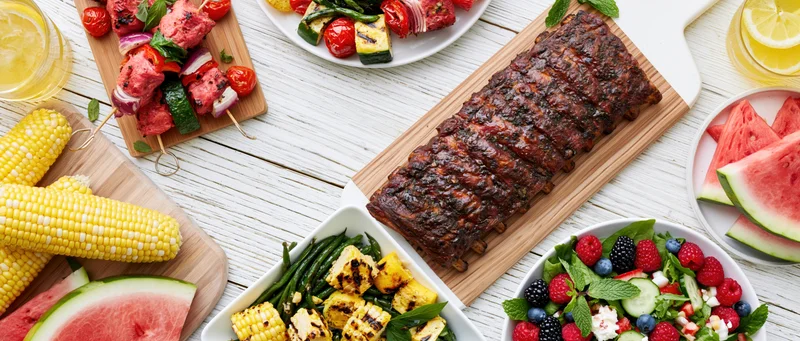 Grilled ribs and summer vegetables arranged on a white wooden table with corn, watermelon, and salad in a double border frame