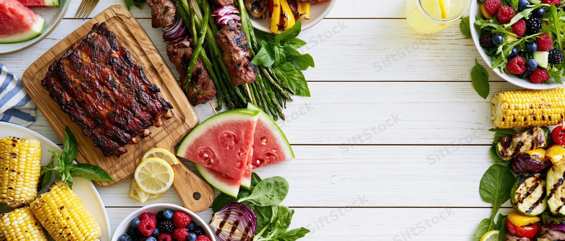 Summer barbecue spread arranged as double border frame on white wooden table with grilled ribs, corn, watermelon, salad, and vegetables