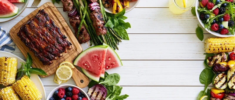 Summer barbecue spread arranged as double border frame on white wooden table with grilled ribs, corn, watermelon, salad, and vegetables