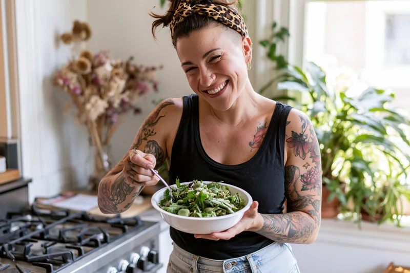 Tattooed woman laughing while holding a bowl of salad in a kitchen
