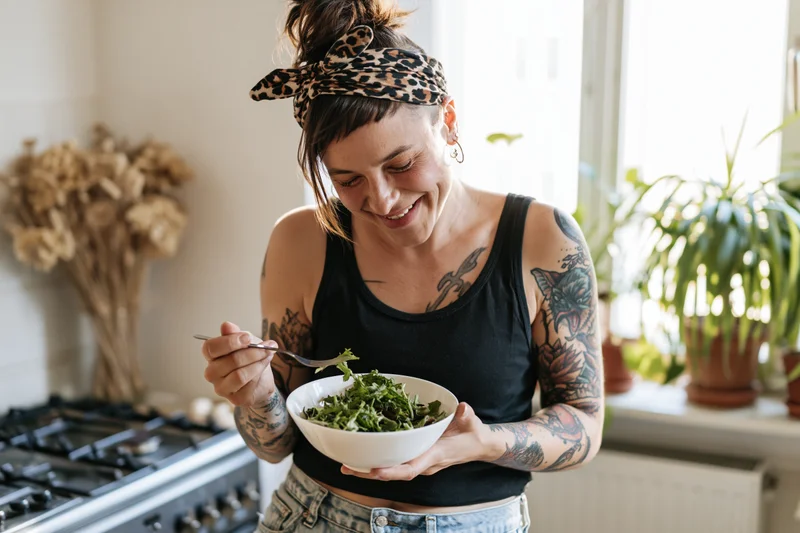 Tattooed young woman laughing while eating a green salad in her kitchen