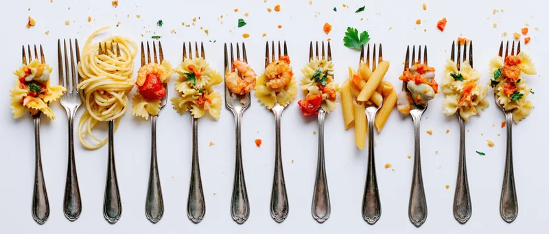 Ten vintage silver forks arranged in a row each holding different pasta dishes on a white background