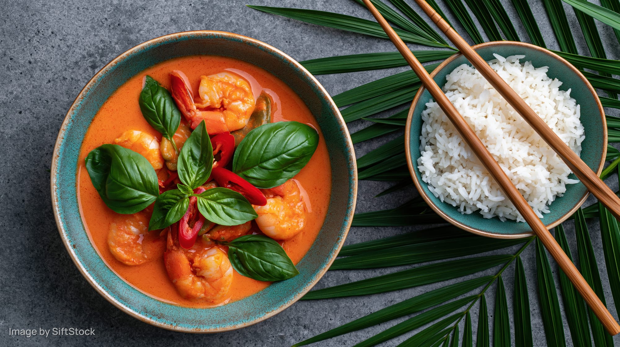 Thai red curry with shrimp in a rustic ceramic bowl garnished with basil and chili, served with rice and chopsticks on a gray surface with palm leaves