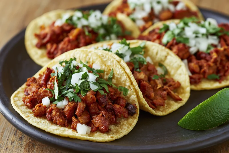 Al pastor tacos on small corn tortillas topped with diced onion and cilantro on a black plate with a lime wedge