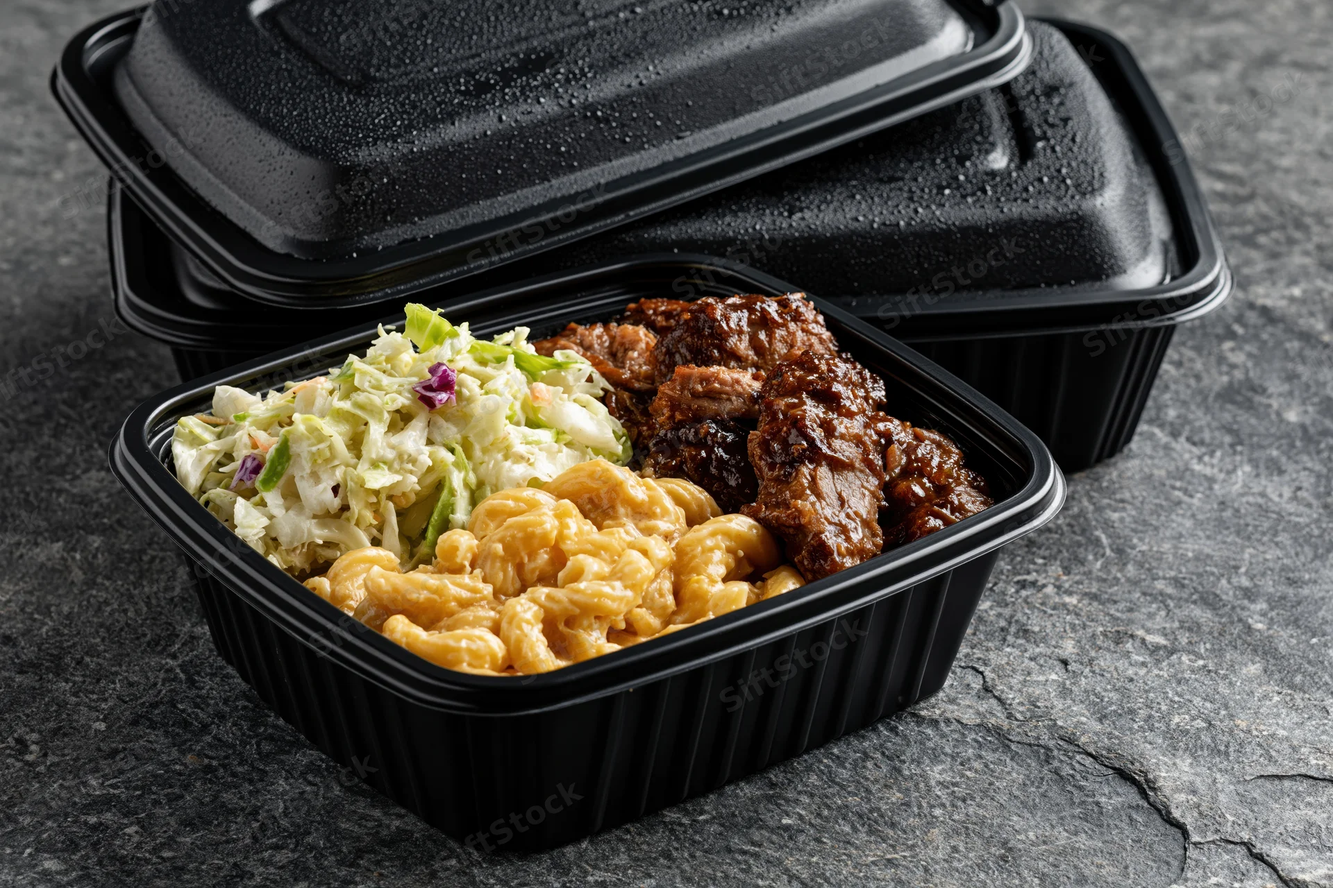 Black plastic meal prep container filled with barbecue ribs, macaroni and cheese, and coleslaw on dark stone surface