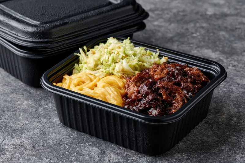 Black plastic meal prep container with pulled pork, macaroni and cheese, and coleslaw on dark stone surface