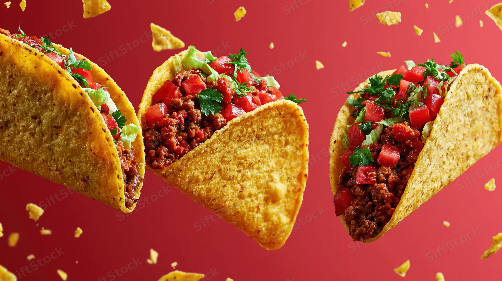Three crispy corn tacos filled with seasoned ground beef and toppings floating in midair against red background