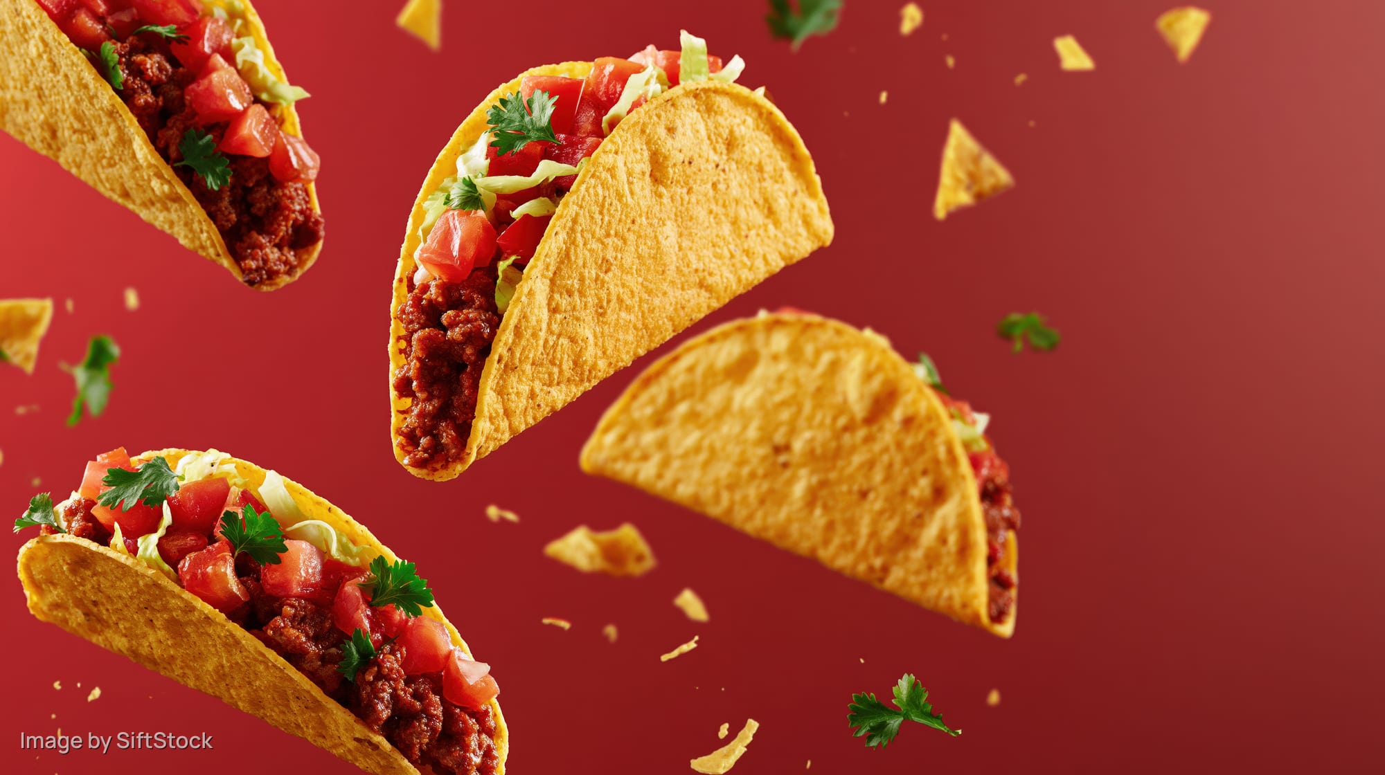 Crispy corn tacos filled with seasoned beef and toppings floating in midair against a red background
