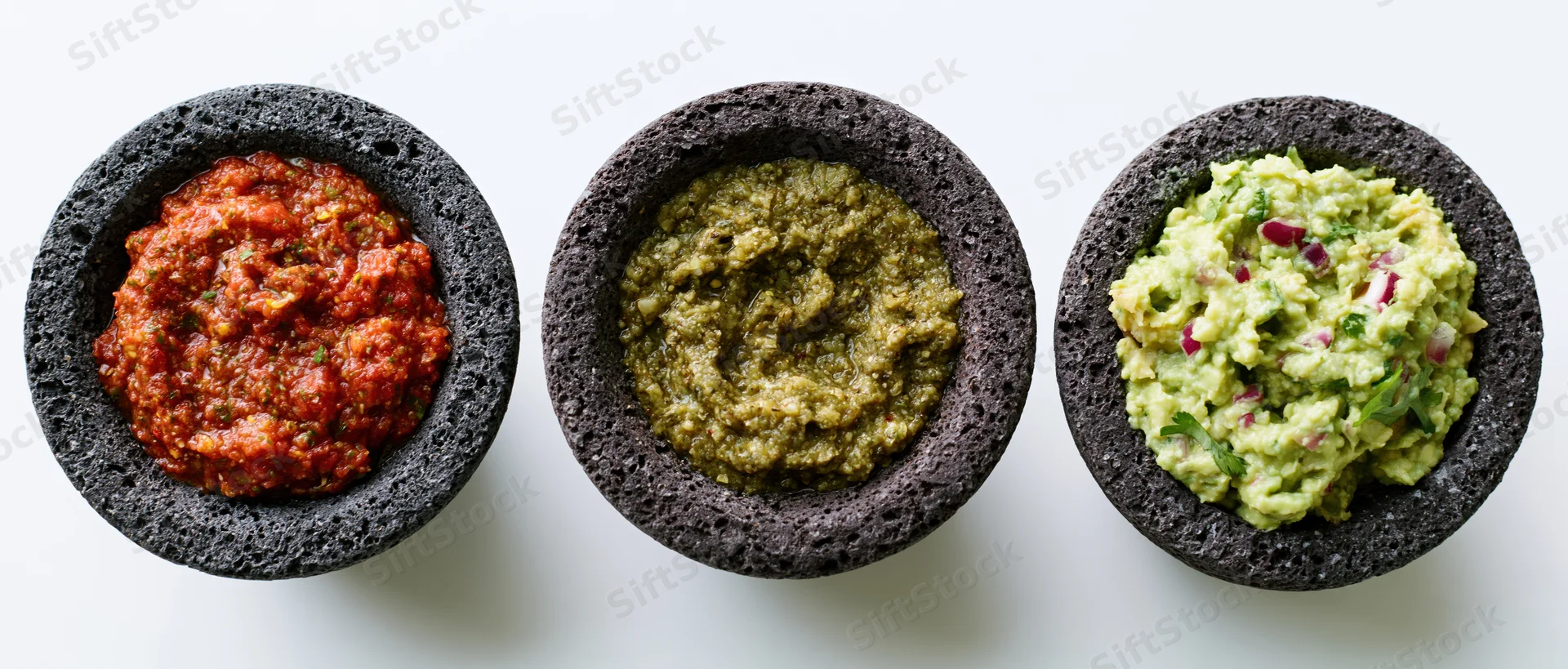 Three dark grey volcanic stone molcajetes each holding a different mexican sauce arranged in a row on a white surface