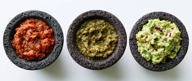 Three dark grey volcanic stone molcajetes each holding a different mexican sauce arranged in a row on a white surface