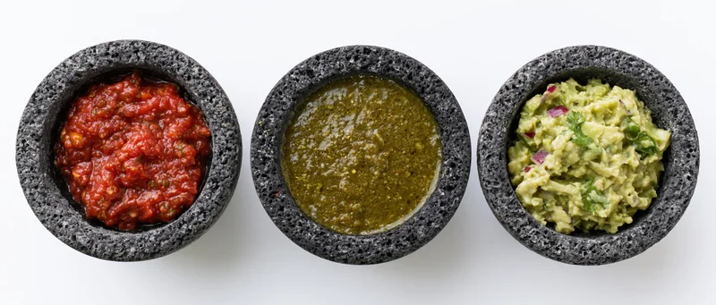 Three dark gray volcanic stone molcajetes each holding a different salsa arranged in a row on a white surface