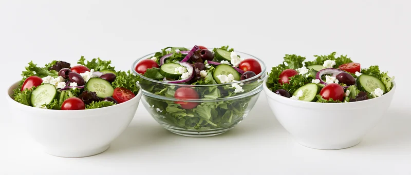 Three fresh garden salads in white and glass bowls arranged side by side on a white surface