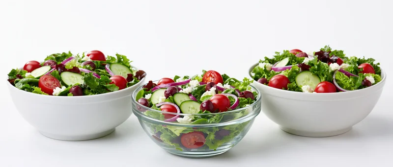Three fresh garden salads in white and glass bowls arranged side by side on a white surface