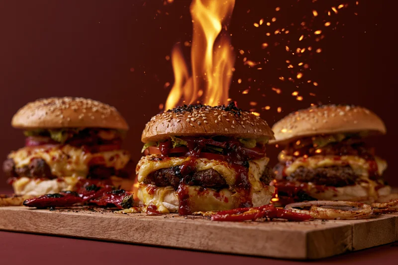 Three loaded beef burgers with cheese lettuce tomato on wooden board with flames and sparks