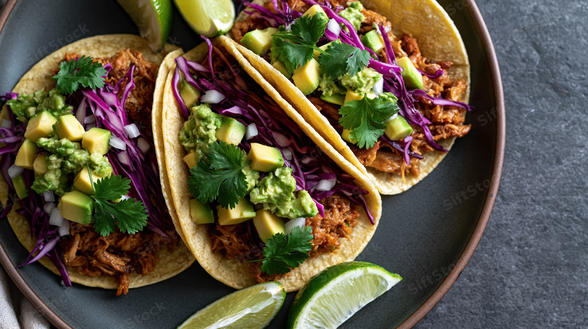 Three pulled pork street tacos in soft corn tortillas topped with guacamole, purple cabbage, cilantro and lime wedges on a dark plate