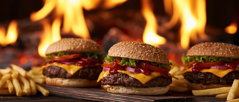 Three cheeseburgers with sesame seed buns and fries on a wooden board in front of flames
