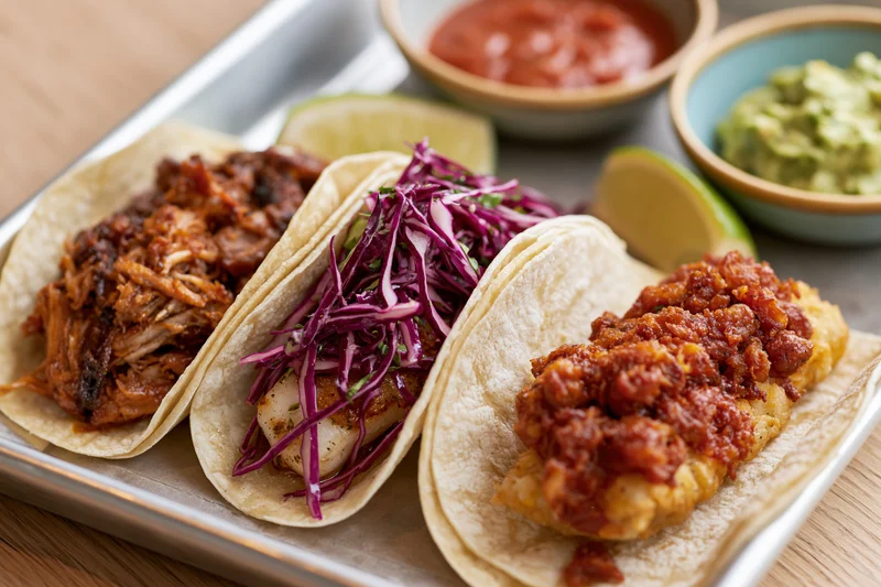 Three street-style tacos with pulled pork, fish, and cabbage on a metal tray with salsa and guacamole on the side