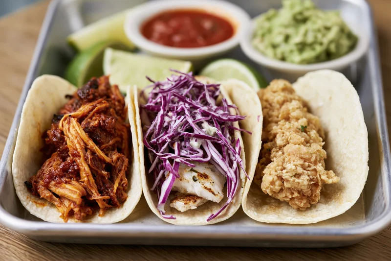 Three street-style tacos with pulled pork, fish, and fried chicken on a metal tray with salsa, guacamole, and lime wedges