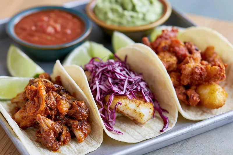 Three street-style tacos with chicken, fish, and pork fillings on a metal tray with salsa, guacamole, and lime wedges