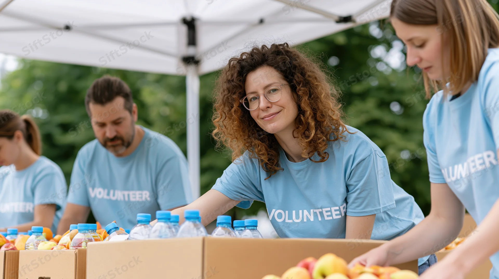 Woman in blue volunteer t-shirt smiling while sorting food donations under a tent with other volunteers