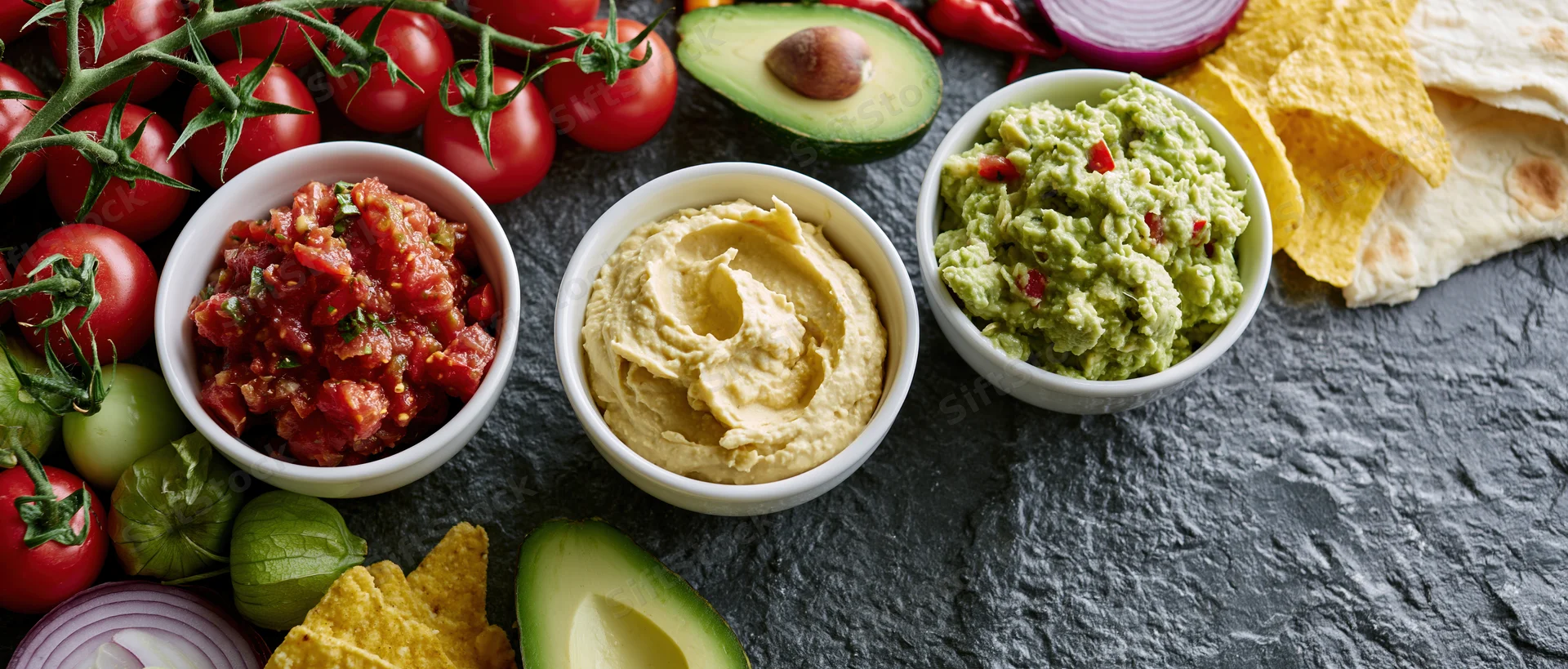 Three white bowls of salsa, hummus and guacamole arranged on a dark slate surface with fresh vegetables and chips around