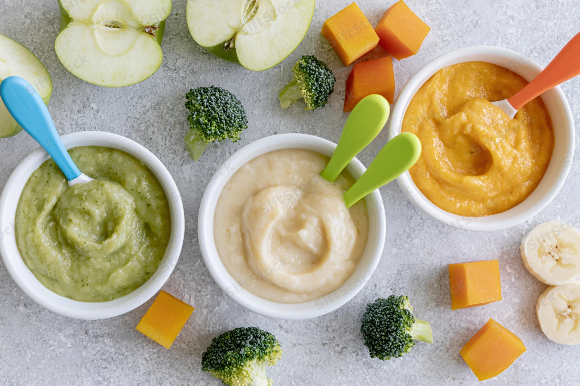 Three white ceramic bowls of baby food puree in green, orange and beige with fresh fruits and vegetables arranged on a countertop