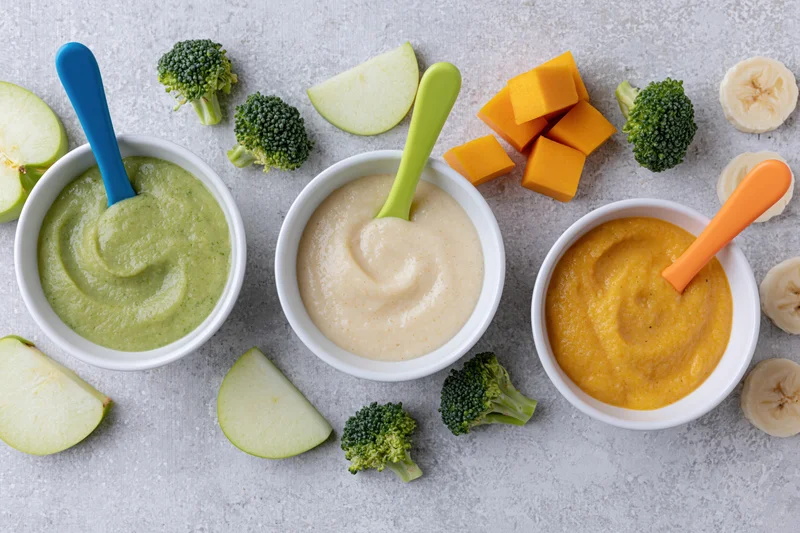 Three white bowls of baby food puree with broccoli, apple, butternut squash and banana arranged on a countertop