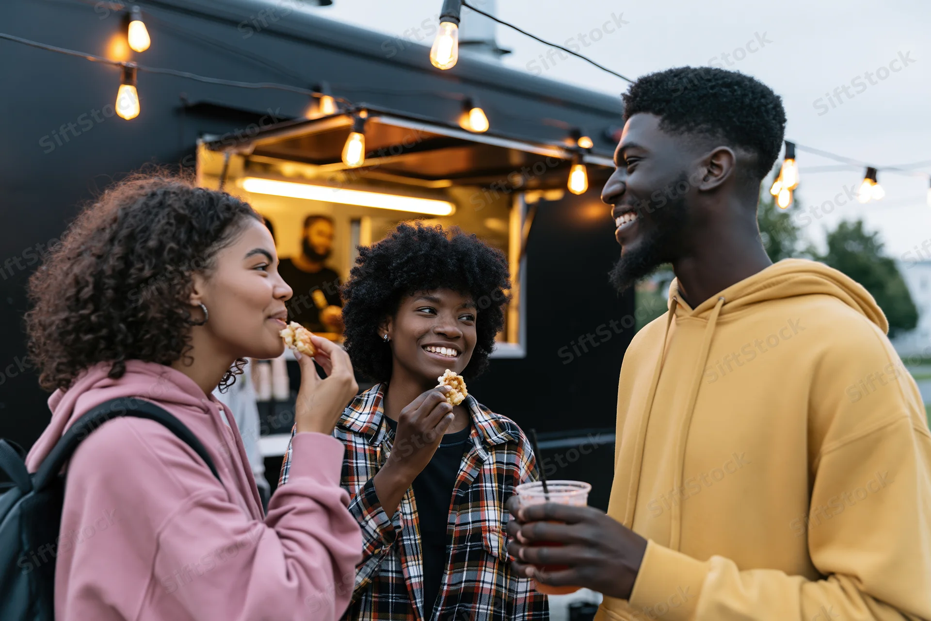 Three young adults eating and drinking together in front of a food truck at evening