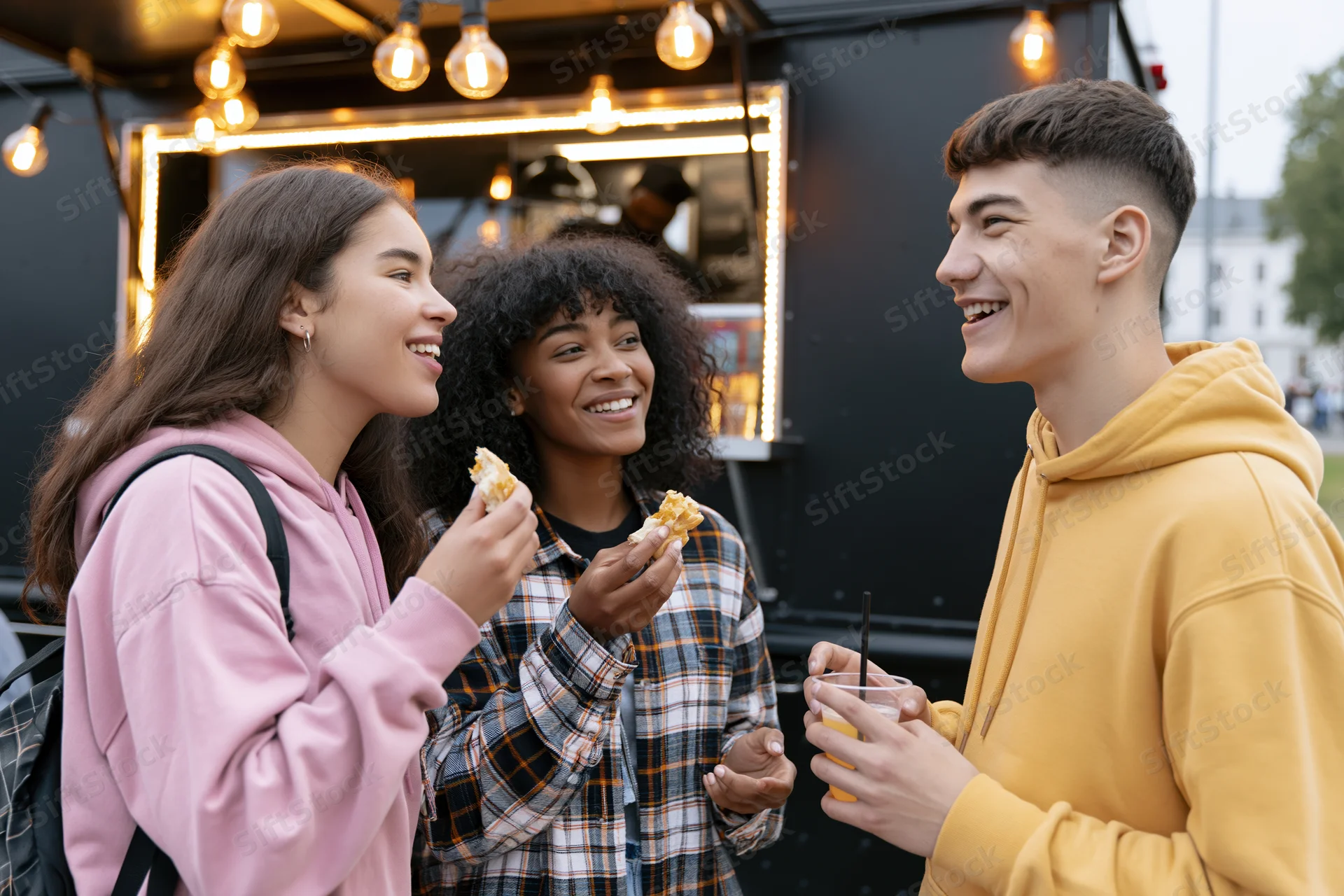 Three young adults smiling and eating snacks together in front of a food truck at evening