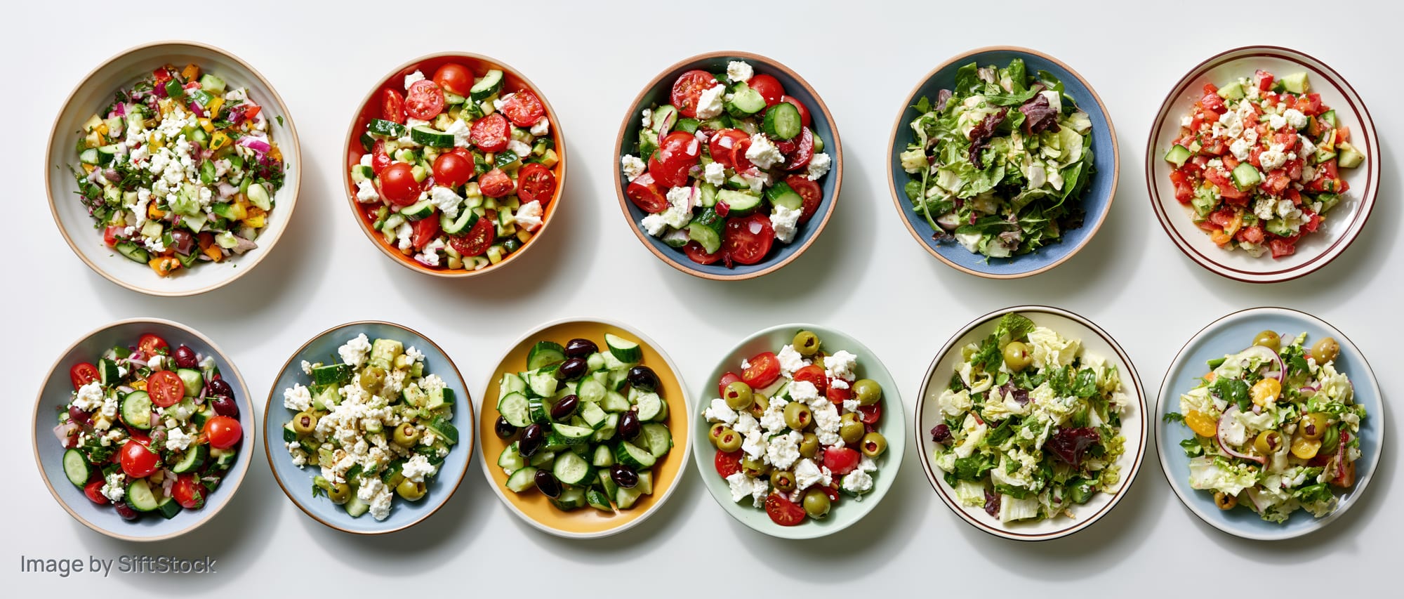 Twelve assorted fresh salads in colorful bowls arranged in two rows on white surface