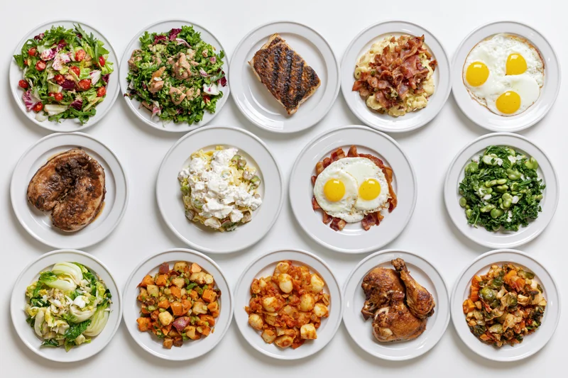 Twelve round plates arranged in grid each holding a different cooked dish viewed from above on white surface