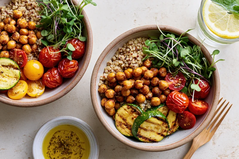 Two ceramic buddha bowls filled with quinoa roasted chickpeas vegetables and microgreens on light surface with fork and glass of lemon water