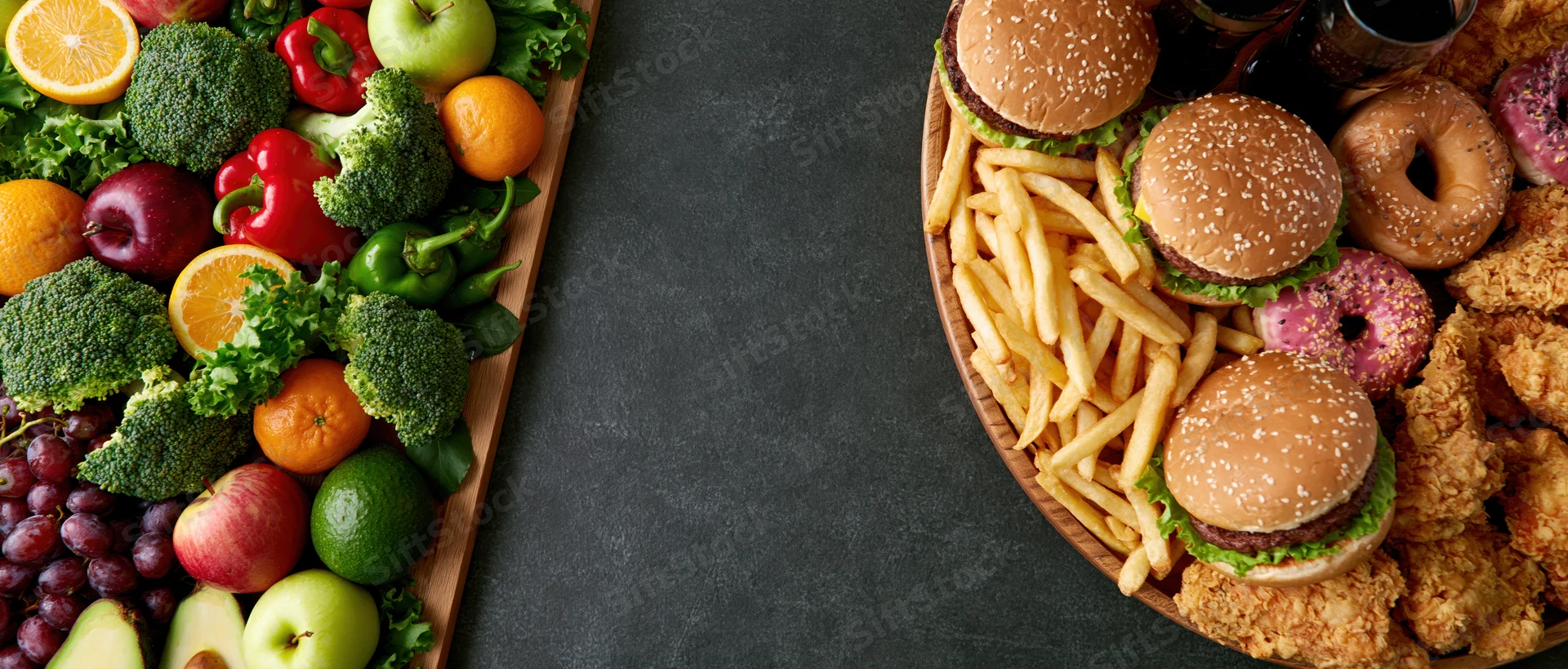 Healthy vegetables and fruits on a wooden tray beside junk food including burgers, fries, donuts and fried chicken on a dark surface