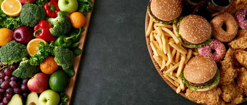 Healthy vegetables and fruits on a wooden tray beside junk food including burgers, fries, donuts and fried chicken on a dark surface