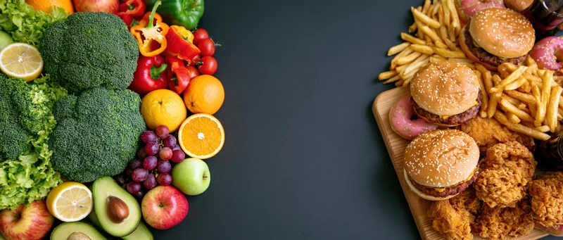 Healthy vegetables and fruits arranged on left side, junk food including burgers and fries on right side, on dark surface with central copyspace