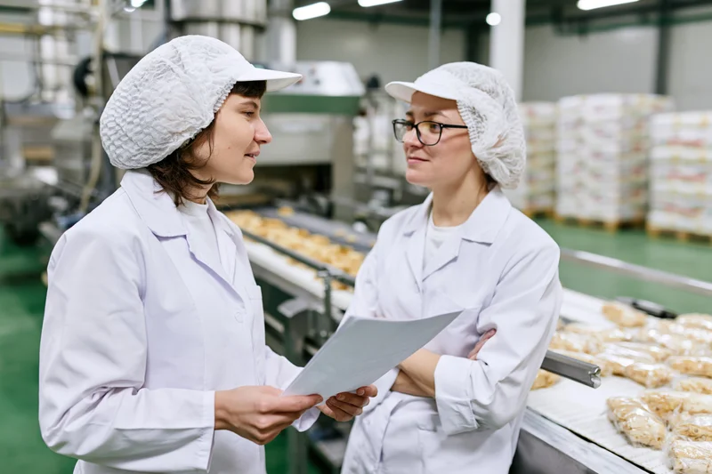 Two women in white lab coats and hairnets discussing a document in a food factory production area