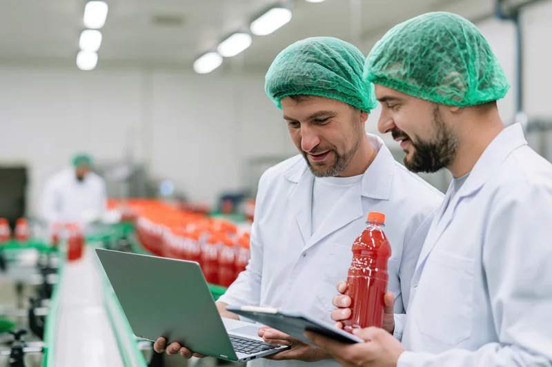 Two men in lab coats and green hairnets inspecting a bottle on a production line while using a laptop and tablet
