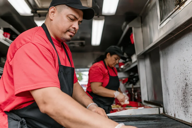 Man and woman cooking on grill in food truck kitchen wearing red shirts and black aprons