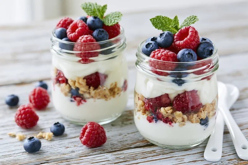 Two glass jars filled with layered yogurt parfait topped with berries and mint on a wooden table