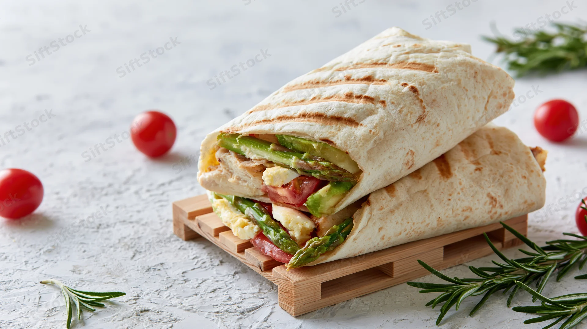 Two grilled tortilla wraps stacked on a small wooden pallet with visible fillings of avocado, asparagus, tomato, egg and chicken on a white textured surface
