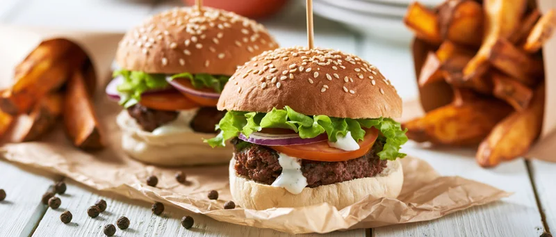 Two beef burgers with sesame buns and toppings served with fries on a wooden table