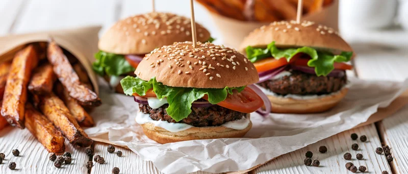 Three beef burgers with sesame buns and fresh vegetables served with fries on parchment paper on a wooden table