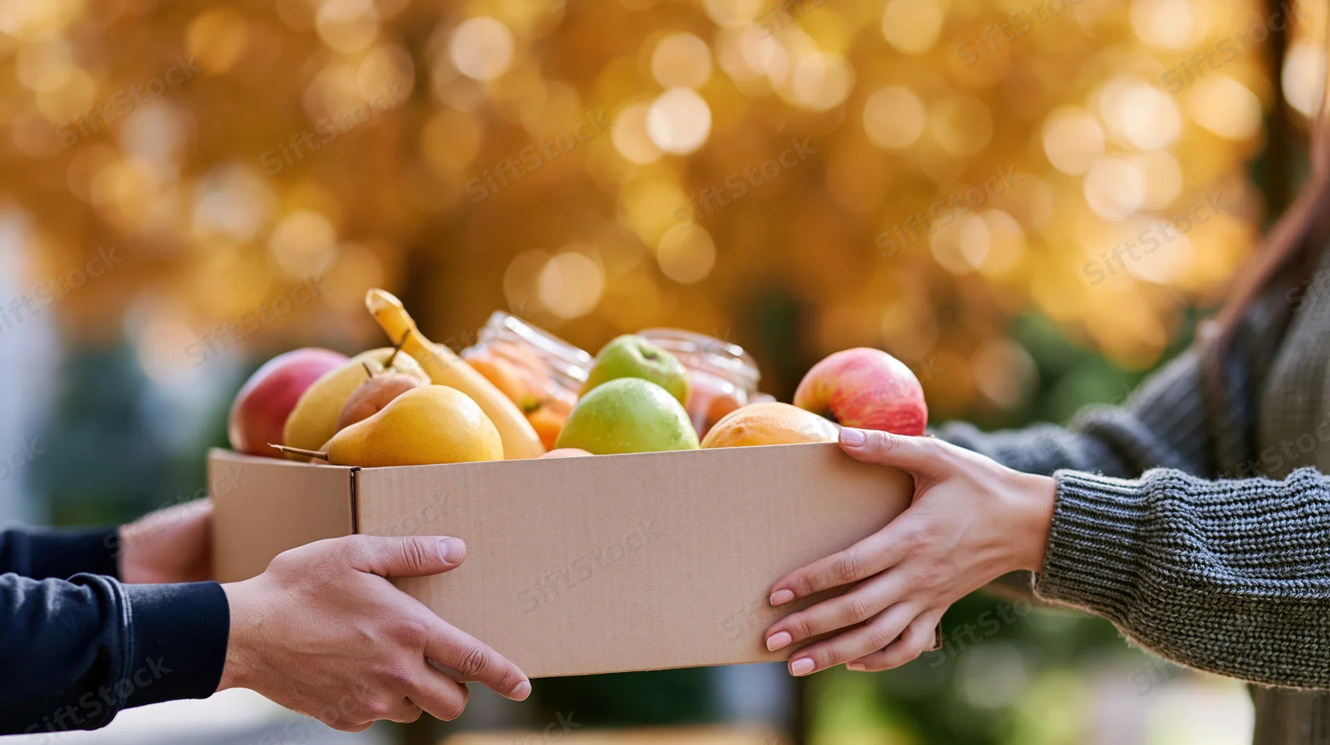 Two people passing a cardboard box filled with fresh fruit outdoors in autumn